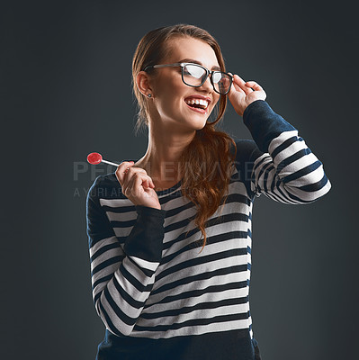 Buy stock photo Studio shot of a cheerful young woman wearing glasses and holding a lollypop while standing against a dark background