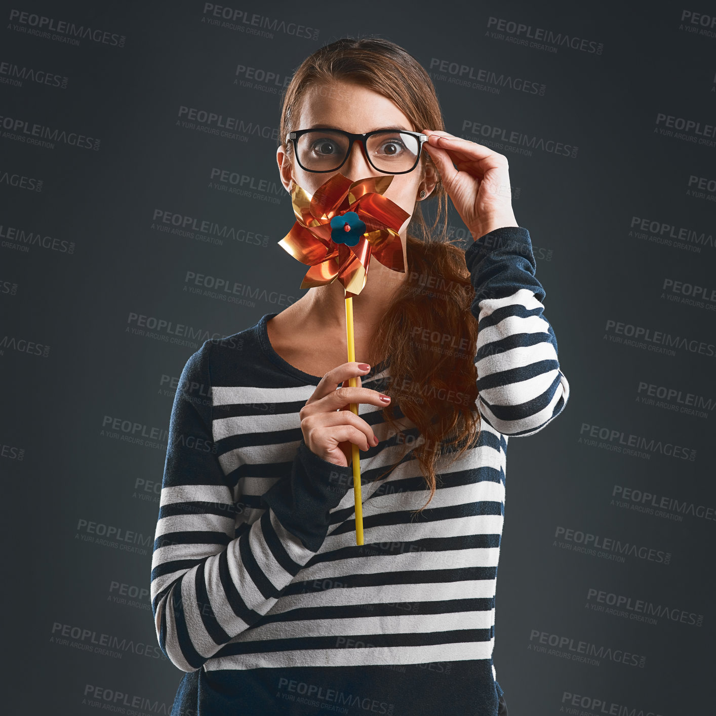 Buy stock photo Studio portrait of an attractive and playful young woman holding a pinwheel and blowing it while standing against a dark background