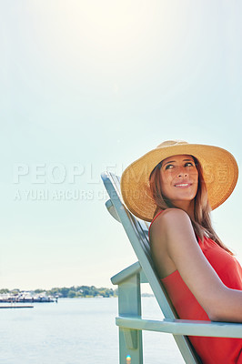 Buy stock photo Shot of a cheerful young woman wearing a hat while being seated on a chair next to a lake outside in the sun