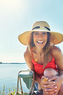 Buy stock photo Portrait of a cheerful young woman wearing a hat while being seated on a chair next to a lake outside in the sun