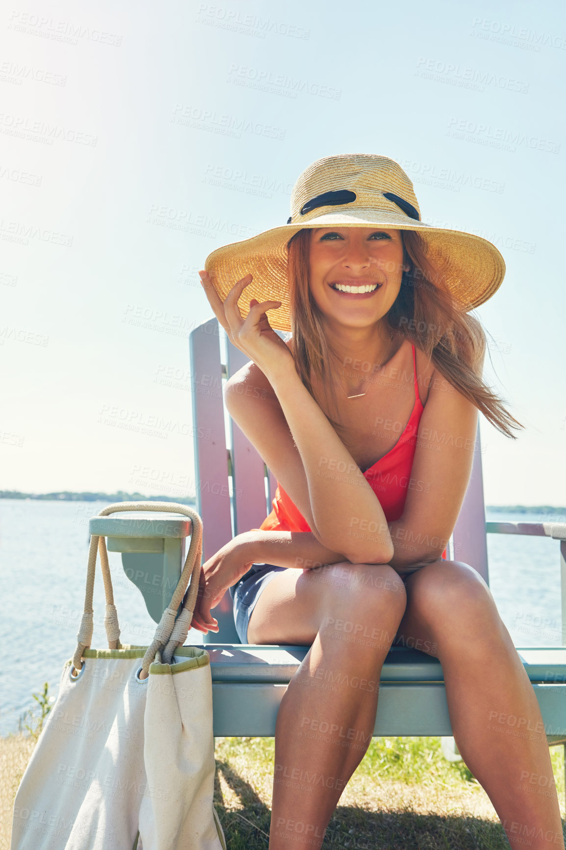 Buy stock photo Portrait of a cheerful young woman wearing a hat while being seated on a chair next to a lake outside in the sun