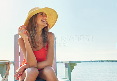 Buy stock photo Shot of a cheerful young woman wearing a hat while being seated on a chair next to a lake outside in the sun