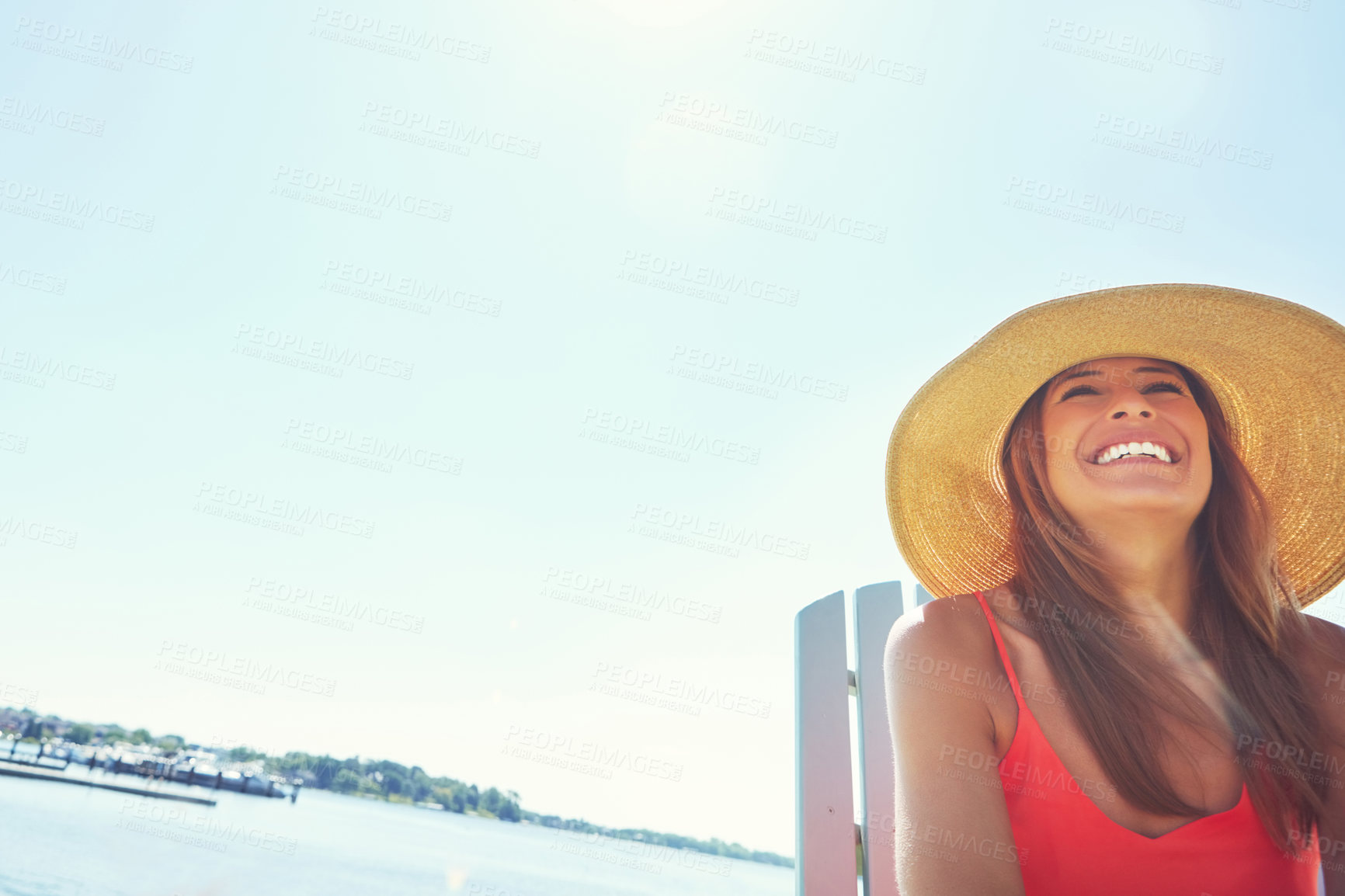 Buy stock photo Shot of a cheerful young woman wearing a hat while being seated on a chair next to a lake outside in the sun