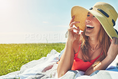 Buy stock photo Shot of a cheerful young woman wearing a hat while lying on a grass field outside in the sun