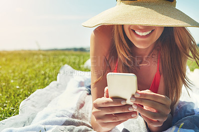 Buy stock photo Shot of a cheerful young woman wearing a hat while lying on the ground busy with her phone outside in the sun