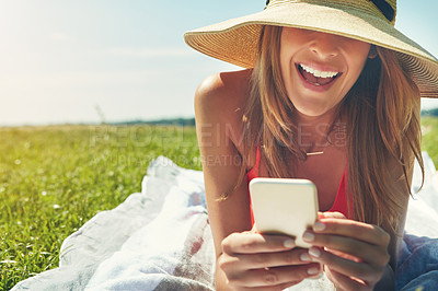 Buy stock photo Shot of a cheerful young woman wearing a hat while lying on the ground busy with her phone outside in the sun