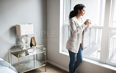 Buy stock photo Shot of a cheerful young woman drinking coffee while looking through a window inside at home during the day