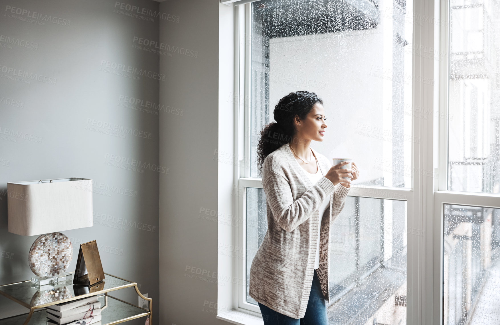 Buy stock photo Shot of a cheerful young woman drinking coffee while looking through a window inside at home during the day