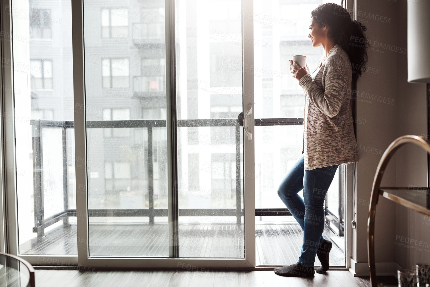 Buy stock photo Shot of a cheerful young woman drinking coffee while looking through a window inside at home during the day