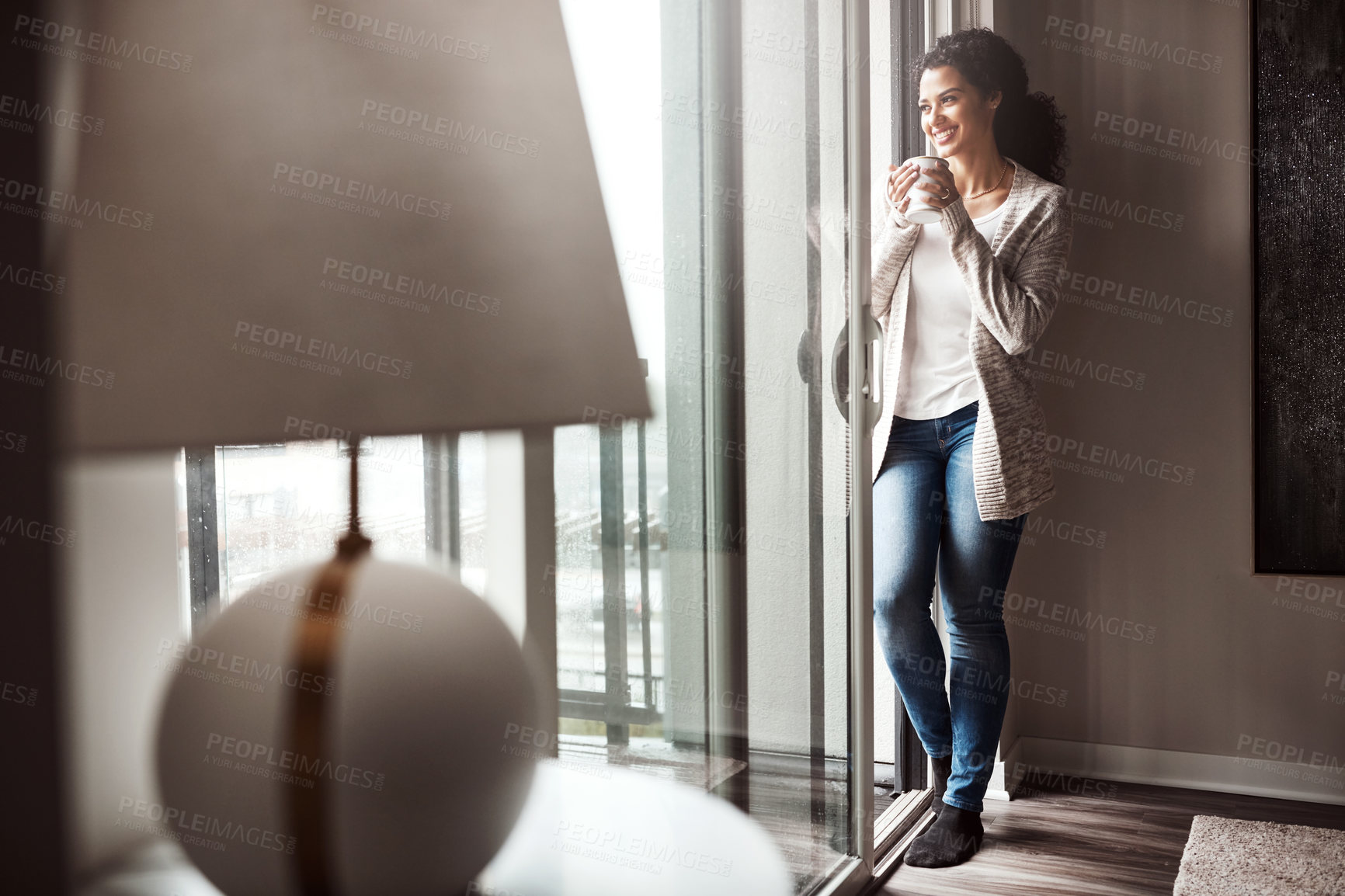 Buy stock photo Shot of a cheerful young woman drinking coffee while looking through a window inside at home during the day