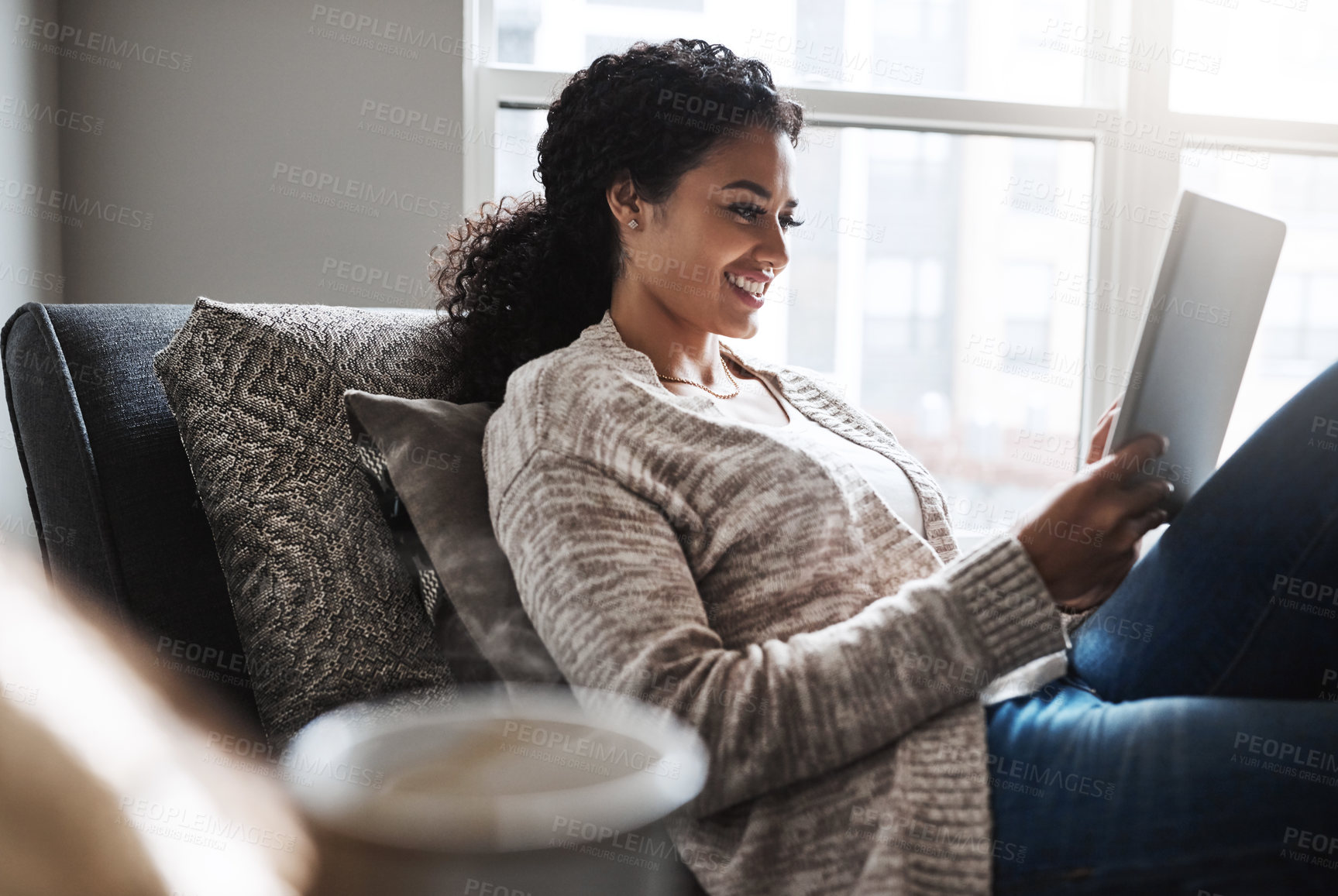 Buy stock photo Shot of a cheerful young woman relaxing on a chair while browsing on a digital tablet at home