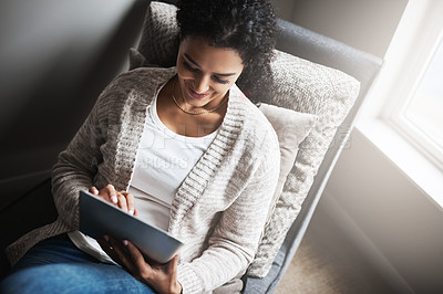 Buy stock photo Shot of a cheerful young woman relaxing on a chair while browsing on a digital tablet at home