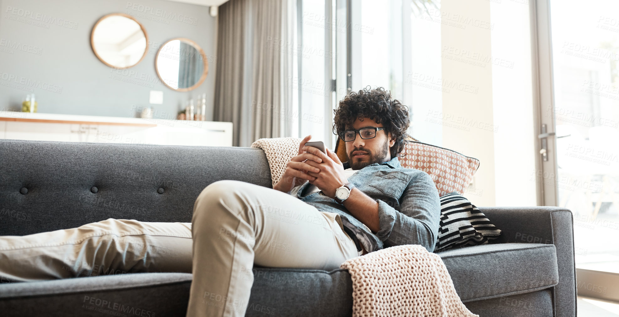 Buy stock photo Shot of a handsome young man using his cellphone while relaxing on the couch at home