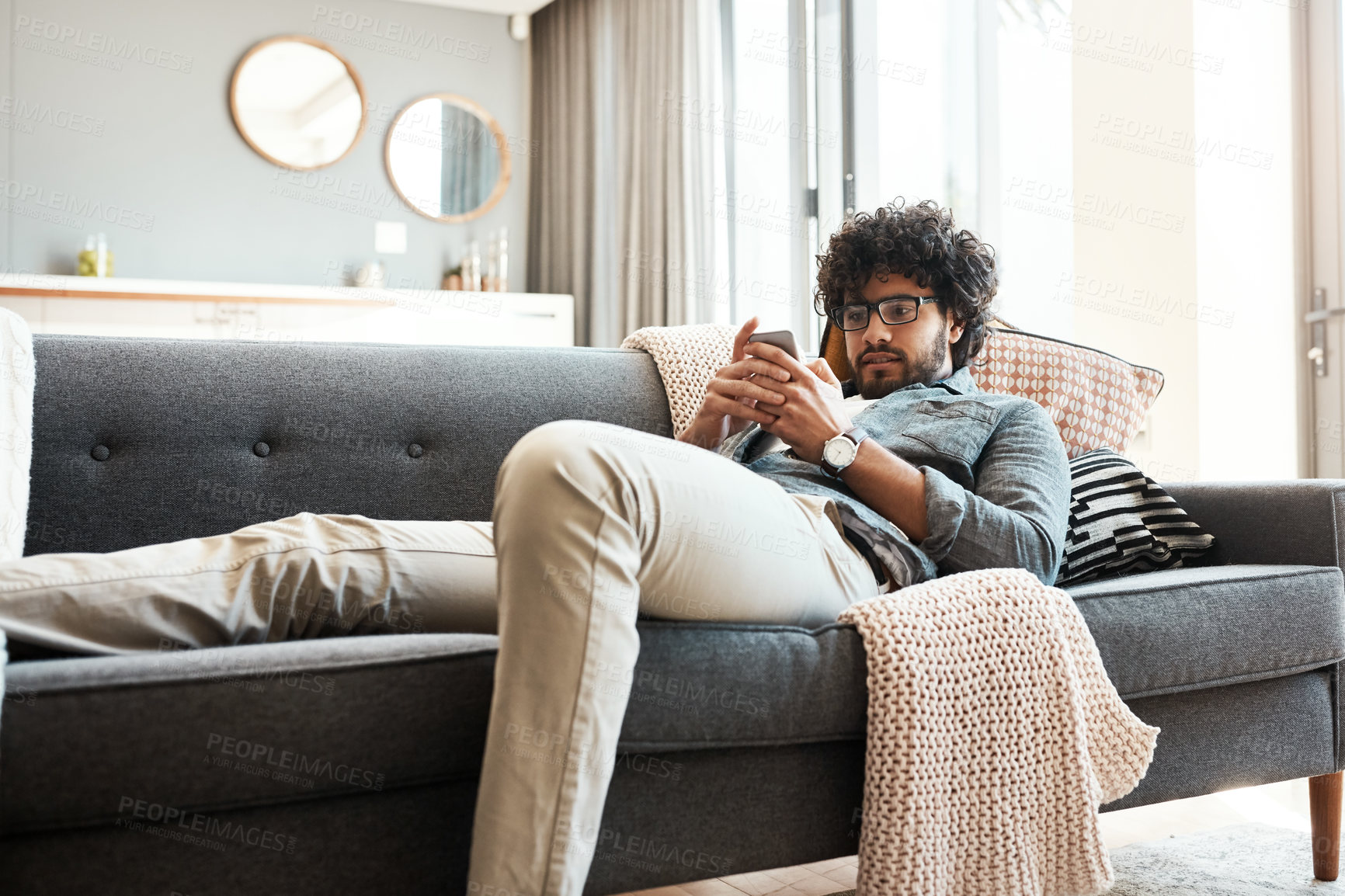 Buy stock photo Shot of a handsome young man using his cellphone while relaxing on the couch at home