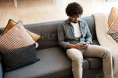 Buy stock photo Shot of a handsome young man using his cellphone while relaxing on the couch at home