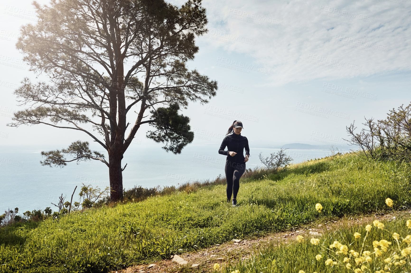 Buy stock photo Shot of a determined young woman running up a hill by herself with a nice view of the ocean in the background