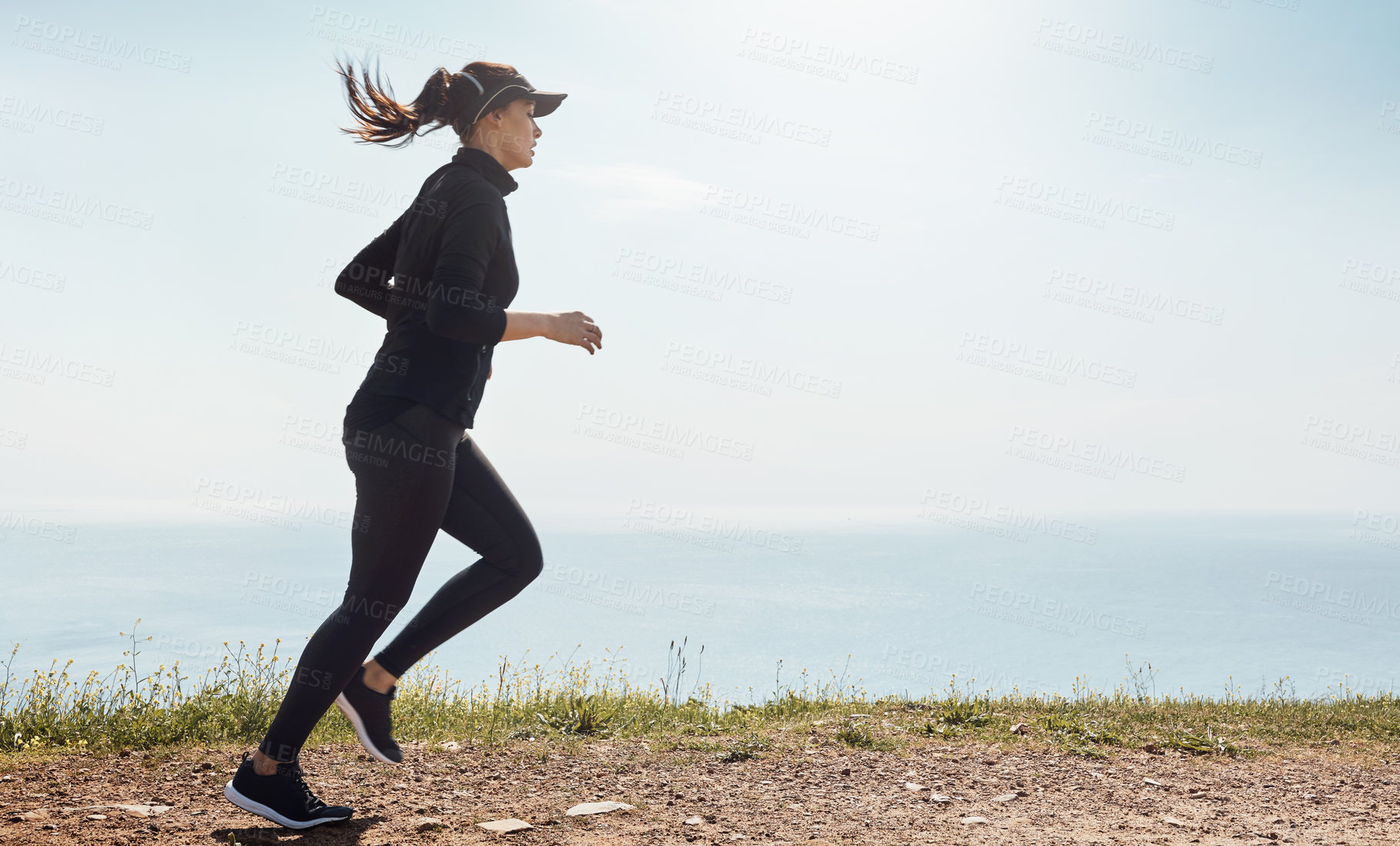 Buy stock photo Shot of a determined young woman going for a jog on her own with a view of the ocean in the background