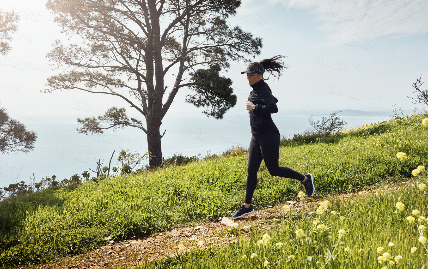 Buy stock photo Shot of a determined young woman running down a hill by herself with a nice view of the ocean in the background