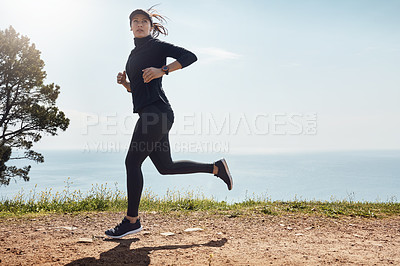 Buy stock photo Shot of a determined young woman going for a jog on her own with a view of the ocean in the background