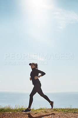 Buy stock photo Shot of a determined young woman going for a jog on her own with a view of the ocean in the background