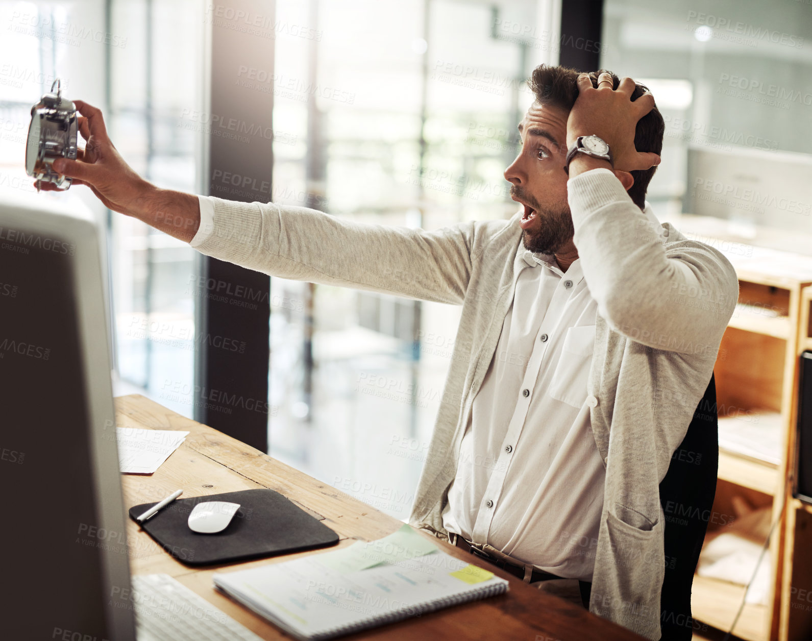 Buy stock photo Shot of a handsome young businessman holding an alarm clock and looking stressed out while working late in an office