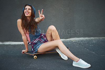 Buy stock photo Portrait of a cheerful young woman seated on a skateboard while showing the peace sign outside during the day