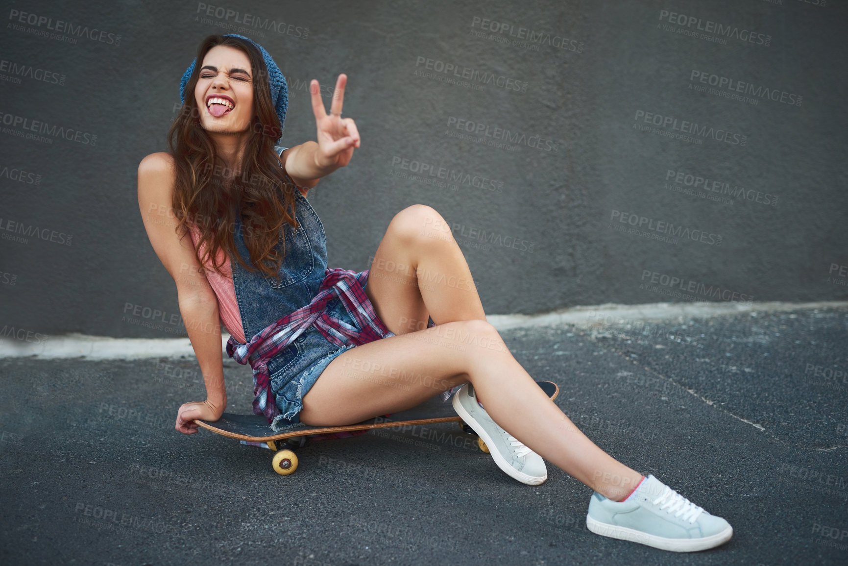 Buy stock photo Portrait of a cheerful young woman seated on a skateboard while showing the peace sign outside during the day