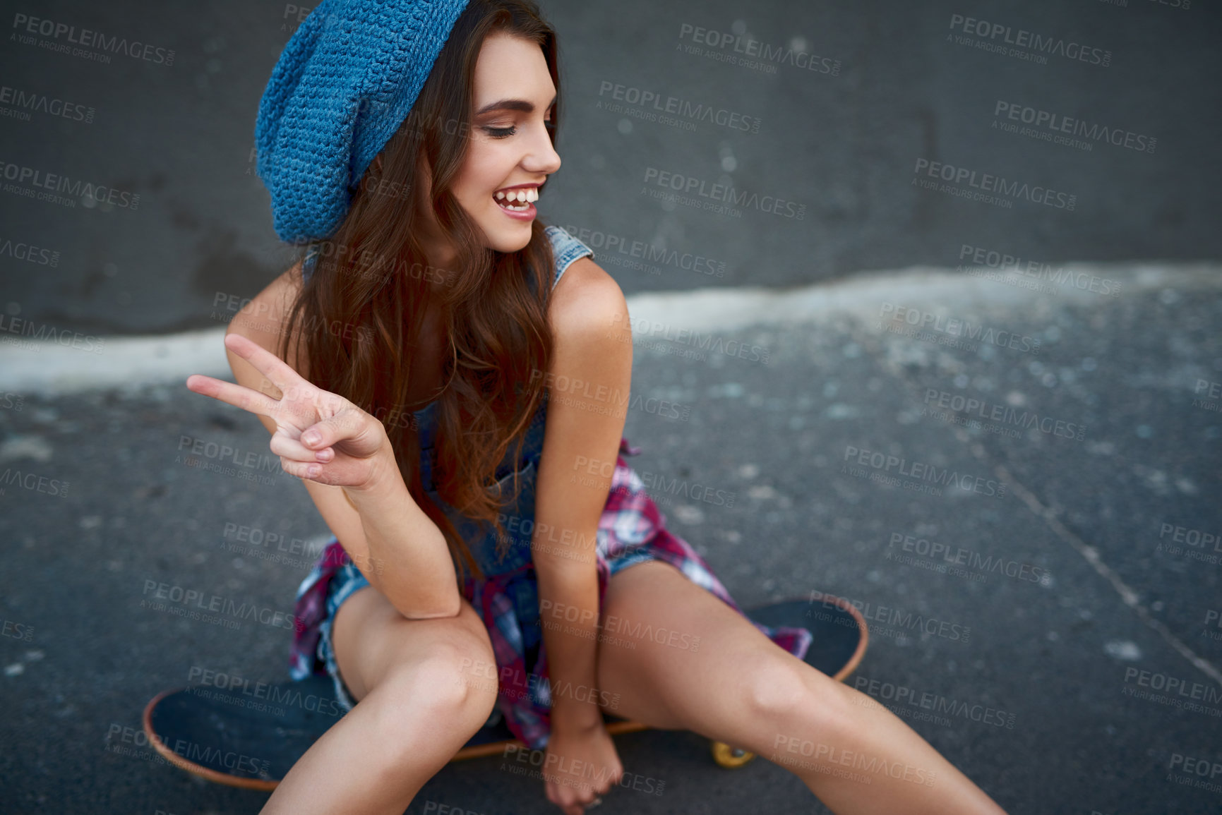 Buy stock photo Shot of a cheerful young woman seated on a skateboard while showing the peace sign outside during the day