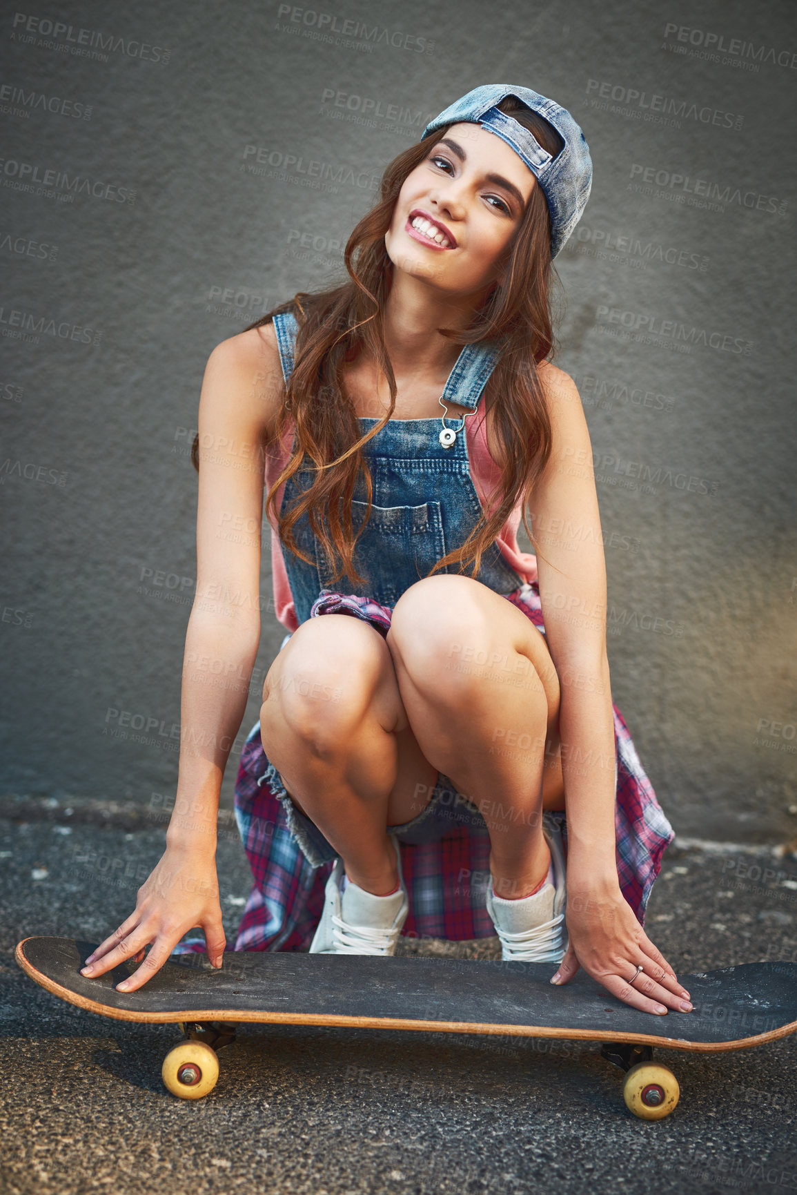 Buy stock photo Portrait of a cheerful young woman wearing a cap while being seated next to a skateboard against a grey background
