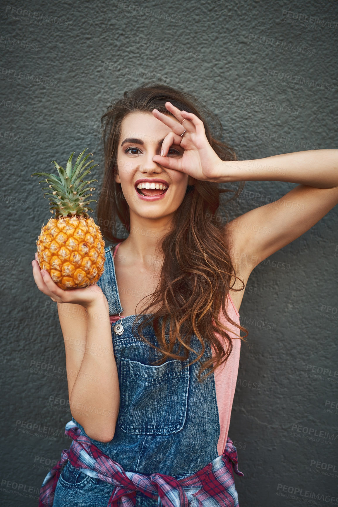 Buy stock photo Portrait of a carefree young woman showing a hand gesture on her face while holding a pineapple against a grey background