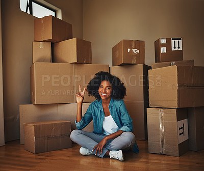 Buy stock photo Portrait of a cheerful young woman seated on the floor while being surrounded by boxes and showing the peace sign in side at home