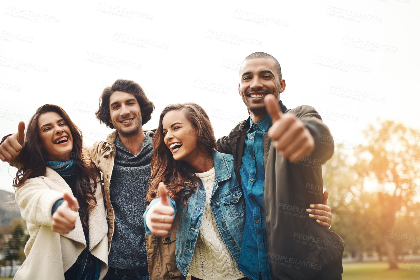 Buy stock photo Portrait of a group of cheerful young friends huddled together while showing thumbs up outside during the day