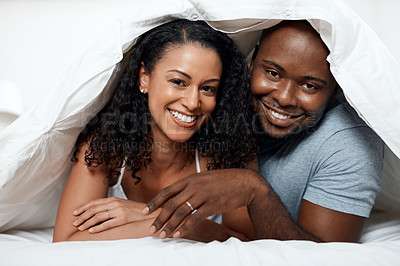 Buy stock photo Portrait of a cheerful young couple lying in bed together under a blanket while looking at the camera at home in the morning