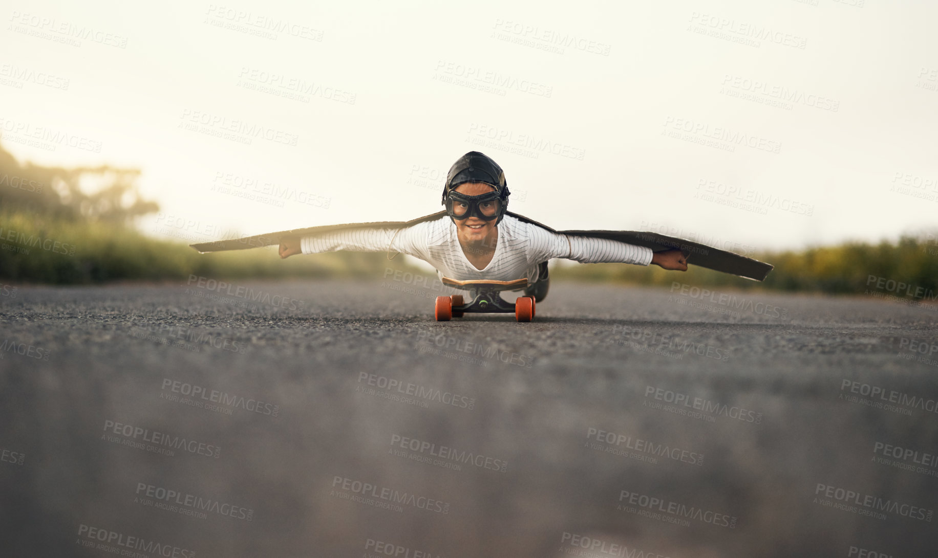 Buy stock photo Portrait of a young boy pretending to fly with a pair of cardboard wings while riding a skateboard outside