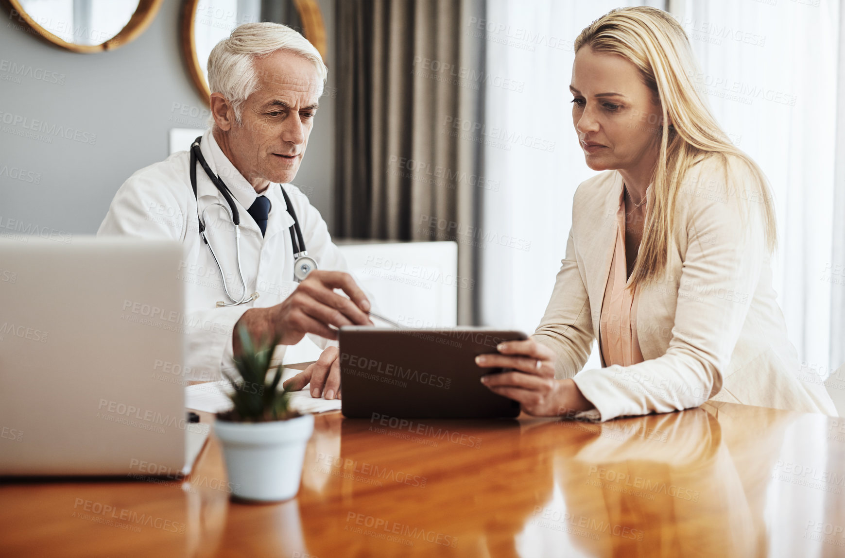 Buy stock photo Shot of a confident mature male doctor consulting with a businesswoman while making use of a digital tablet at a table during the day