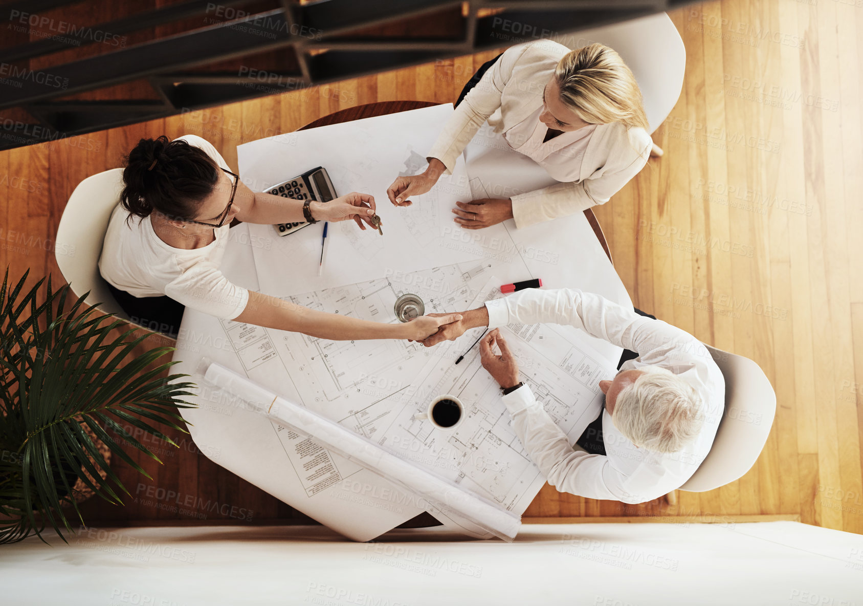 Buy stock photo High angle shot of a group of architects working together on blueprints of a house around a table while shaking hands in agreement inside of a building