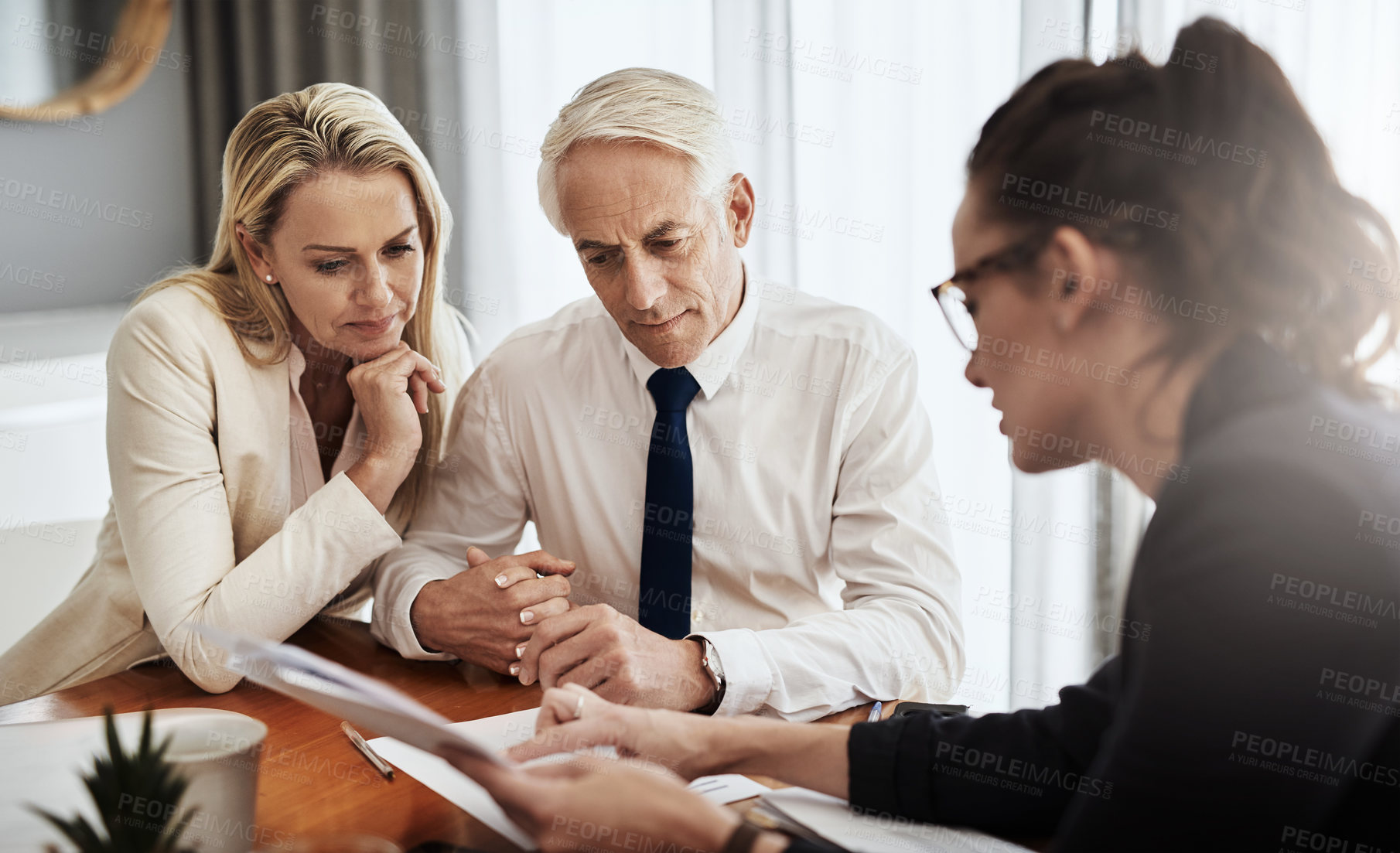 Buy stock photo Shot of a focused mature couple negotiating with a architect over house plans around a table inside of a building during the day