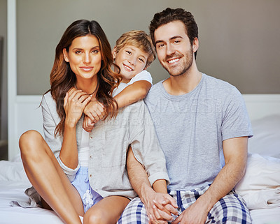 Buy stock photo Portrait of a cheerful young family seated on bed while holding each other and looking at the camera at home in the morning