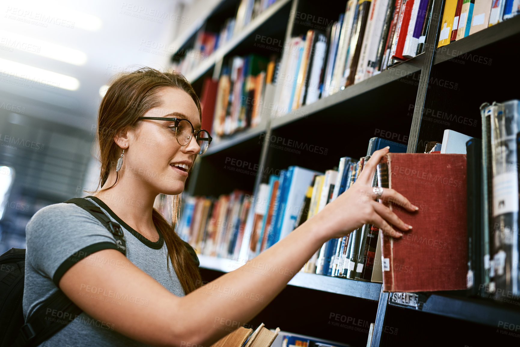 Buy stock photo Shot of a happy young woman removing a book from a shelf in a college library