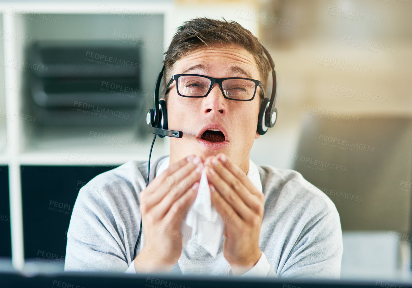 Buy stock photo Shot of a young call centre agent sneezing while working in an office