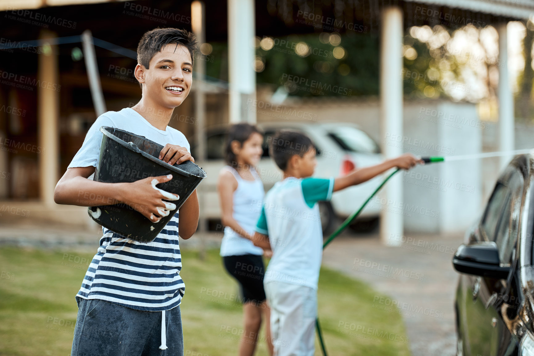 Buy stock photo Boy child and portrait for washing car, outdoor and siblings with garden hose, foam and bonding with chores. Kids, water and bucket with cleaning, soap or smile on lawn by family vehicle in Turkey