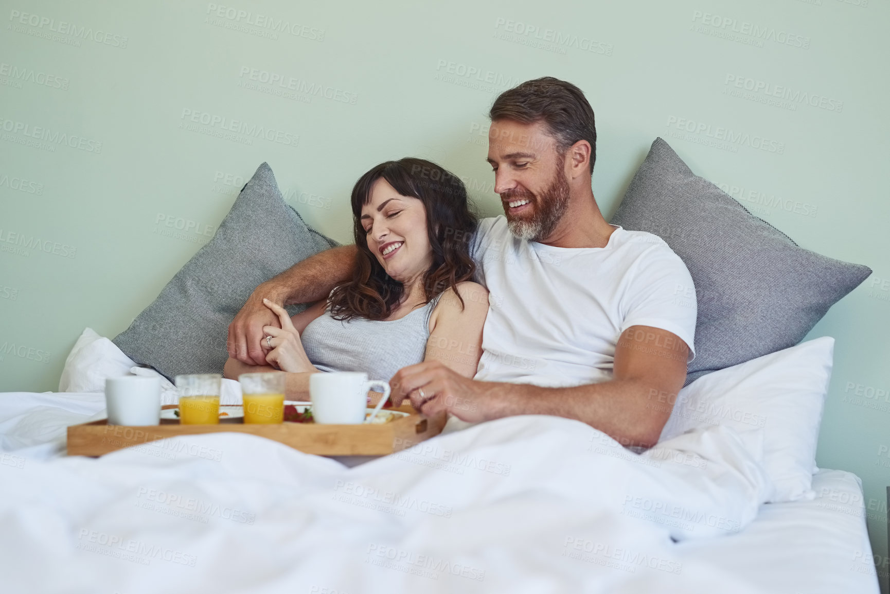 Buy stock photo Shot of a cheerful young couple sitting in bed while enjoying breakfast together during morning hours
