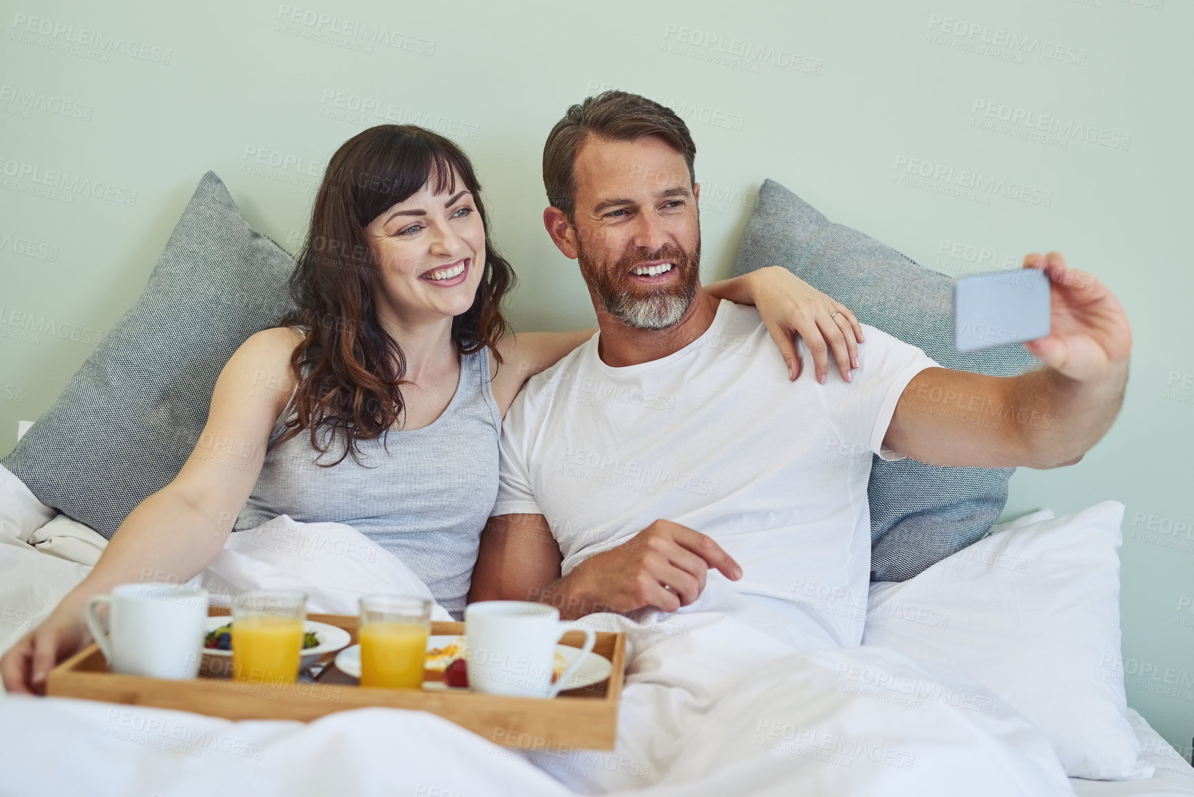 Buy stock photo Shot of a cheerful young couple sitting in bed while enjoying breakfast together and taking a selfie during morning hours