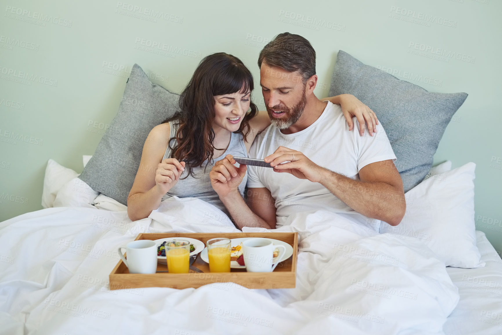 Buy stock photo Shot of a cheerful young couple sitting in bed while enjoying breakfast together and taking a picture during morning hours