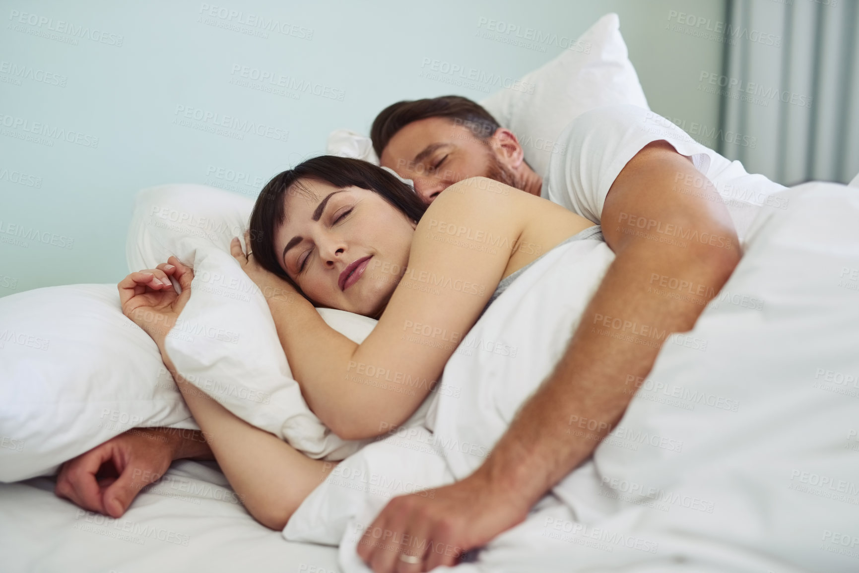 Buy stock photo Shot of a relaxed young couple sleeping in each other's arms in bed during morning hours