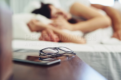 Buy stock photo Shot of a pair of glasses and a cellphone standing next to each other on a bedside table at home during the day
