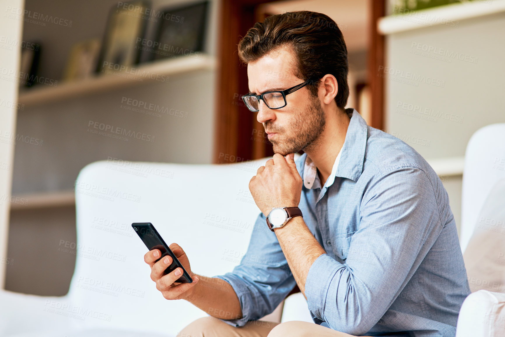 Buy stock photo Shot of a handsome young man looking confused while using a cellphone at home