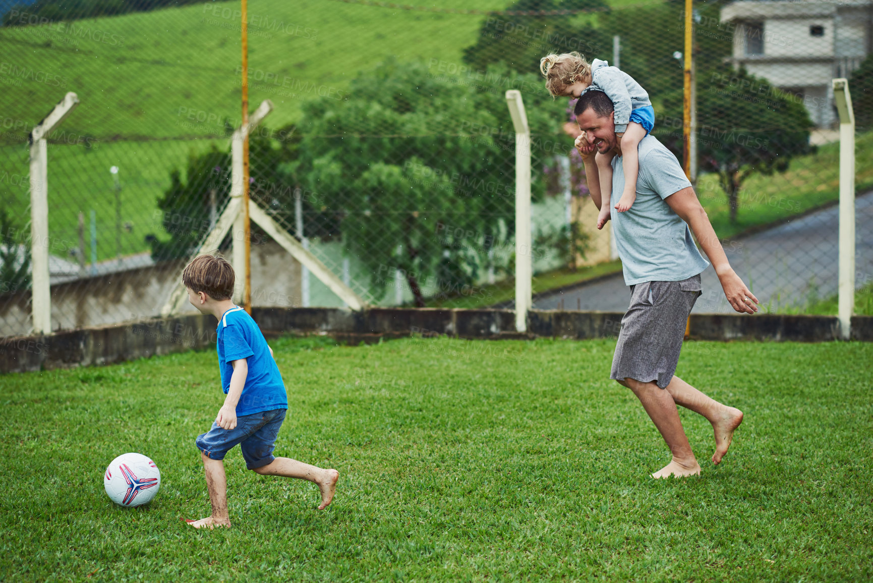 Buy stock photo Shot of a cheerful middle aged man carrying his daughter on his solders while playing soccer with his son outside during a cloudy day