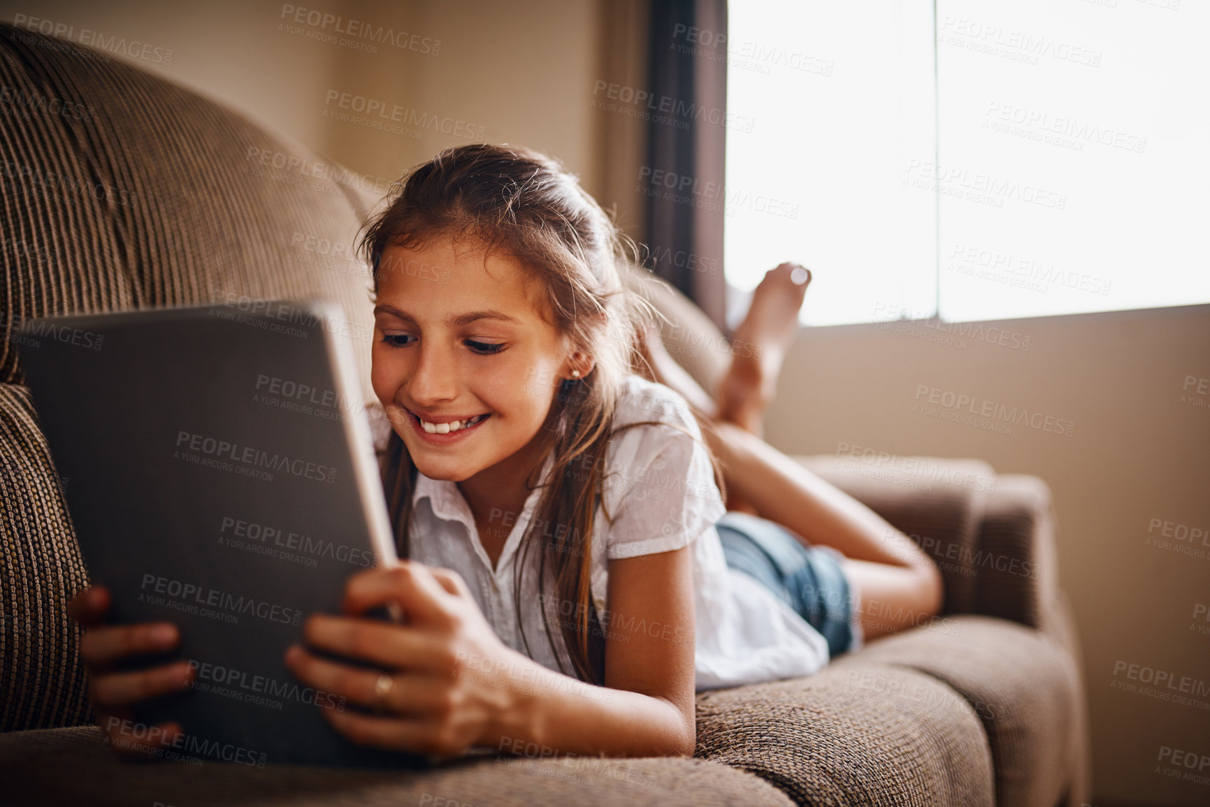 Buy stock photo Full length shot of a young girl using her tablet while lying on a sofa at home