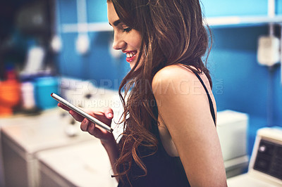 Buy stock photo Shot of an attractive young woman texting on her phone while she does her washing inside of a laundry room during the day